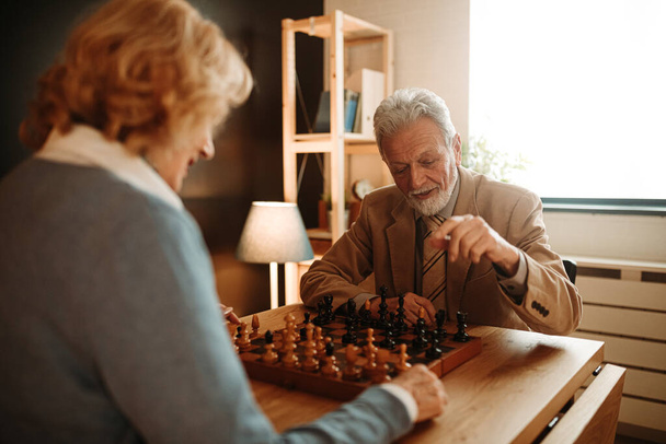 Портрет пожилого человека в костюме и галстуке, играющего в шахматы с женой дома. Он играет с черными фигурами
. - Фото, изображение