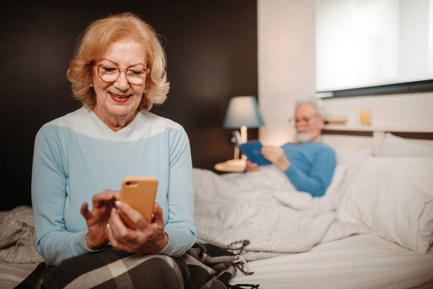 Schließendes Foto einer blonden Frau, die auf ihr Smartphone schaut, während sie im Bett sitzt. Älterer Mann mit grauen Haaren und Bart liegt im Bett und liest ein Buch. - Foto, Bild