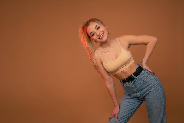 Attractive blonde woman in beige top and blue jeans leaned on her leg smiling and looking at the camera isolated over orange background - Zdjęcie, obraz
