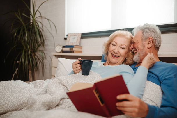Elderly woman is drinking coffee while her husband is reading book. Senior couple enjoys every second of their time while lying in bed. - Photo, image
