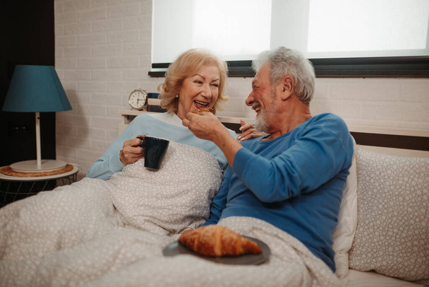 Ridere coppia anziana si sta divertendo mentre seduto sul letto in camera da letto. Gli uomini anziani offrono sua moglie con croissant mentre lei beve caffè del mattino
. - Foto, immagini