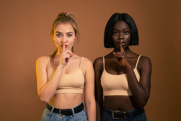 Two beautiful confident young international girls standing isolated over dark orange background, african and european females showing silence gesture - Fotoğraf, Görsel