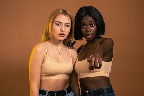 Two beautiful international female, wearing same beige bra, having surprised shocked looks, african girl pointing finger at camera. - Foto, Imagem