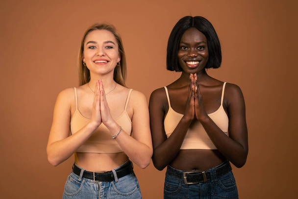 Fiatal, boldog szőke európai nő és afrikai mosolygós lány jógázik, fogja a kezét Namaste-ban és csukva tartja a szemét. A nők otthon meditálnak, békéért és szeretetért imádkoznak, nyugodtak. - Fotó, kép