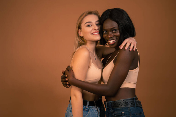 Feliz sorrindo casal lésbico atraente internacional em sutiã bege, meninas europeias e africanas abraçando e olhando para a câmera isolada sobre fundo laranja escuro
 - Foto, Imagem