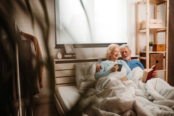 Pareja mayor en casa. Hombre de edad avanzada bien parecido y su esposa de edad avanzada bien parecido están disfrutando de pasar tiempo juntos mientras están acostados en la cama
. - Foto, Imagen