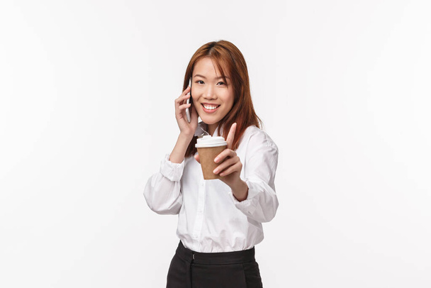 Travail de bureau, personnes et concept de style de vie. Joyeux stupide jeune femme asiatique vous suggérant sa tasse de café tout en parlant au téléphone, appareil photo souriant joyeux, avoir la conversation, fond blanc
 - Photo, image