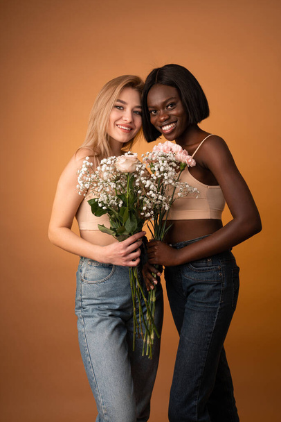 Happy smiling girls in blue jeans and beige top looking at the camera, european and african girls holding flowers - Fotografie, Obrázek