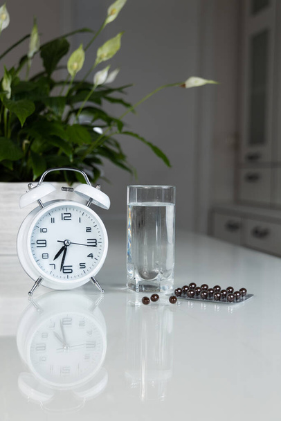 White alarm clock and medical pills with a glass of water on the table. Healthcare and medicine. Vitamins and mineral supplements in capsules. Vertical shot. - Fotografie, Obrázek