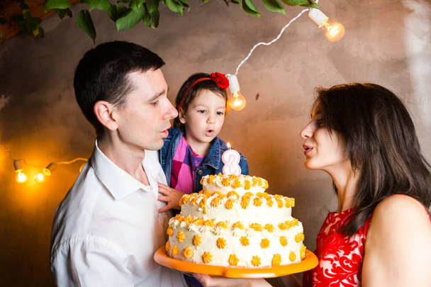 Tema fiesta familiar cumpleaños de los niños y soplar velas en la torta grande. familia joven de tres personas de pie y la celebración de 5 años de edad hija en el patio contra la pared gris y guirnalda bulbos amarillos
. - Foto, Imagen