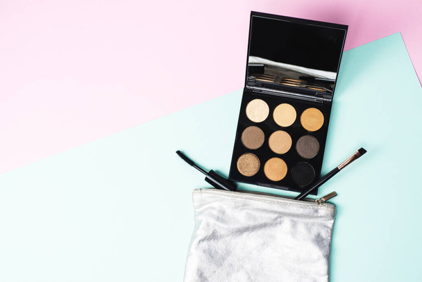A small silver cosmetic bag with a large black palette of neutral neutral tones, a mascara brush and a brush for applying shadows on a blue and pink background. The concept of nude daily makeup. Make up at work. - Photo, Image