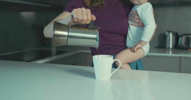A young mother is in the kitchen holding her toddler and pouring a cup of coffee - Filmati, video