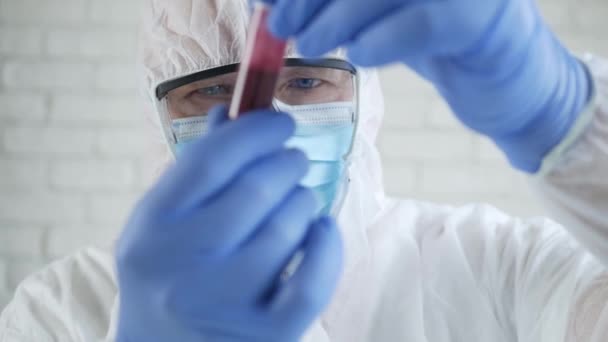 Lekarz w szpitalnym laboratorium noszący kombinezon ochronny Patrząc na rurkę z krwią zainfekowaną koronawirusem - Materiał filmowy, wideo
