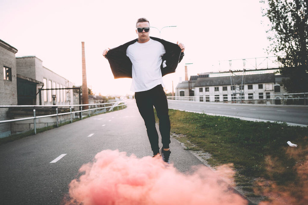 Man in a leather jacket standing at the street with smoke bomb. Colorful portrait of european teenager with red smoke bomb - Fotografie, Obrázek