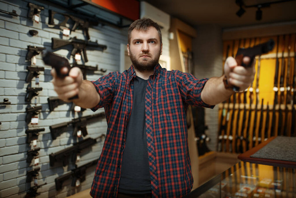 Ο άνθρωπος σημαδεύει με δύο πιστόλια στο οπλοπωλείο. Άνδρας που αγοράζει πιστόλι για ασφάλεια σε κατάστημα όπλων, αυτοάμυνα και σκοποβολή χόμπι - Φωτογραφία, εικόνα