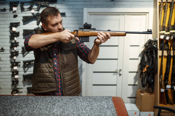 Man aims with new rifle in gun shop. Male person buying weapon in store, hunting and sport shooting hobby - Foto, Imagen