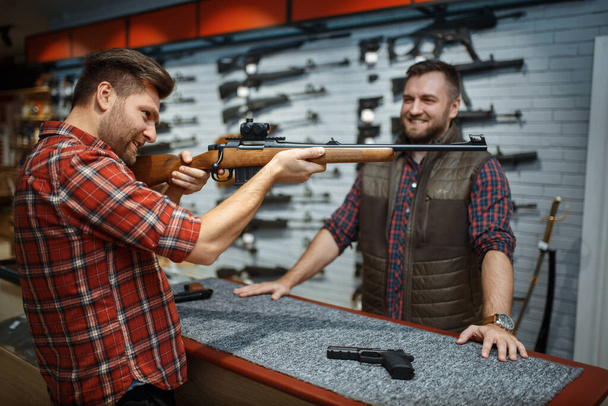 Man aims with new rifle, seller at counter in gun shop. Male person buying weapon in store, hunting and sport shooting hobby - Photo, Image