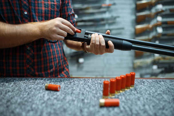 Male person with rifle loads ammo at showcase in gun shop. Euqipment for hunters on stand in weapon store, hunting and sport shooting hobby - Foto, Imagem