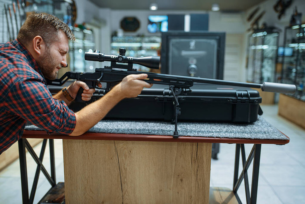 Man aims with sniper rifle in gun shop. Euqipment for hunters on stand in weapon store, hunting and sport shooting hobby  - Photo, Image