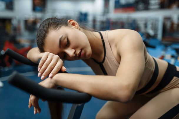 Tired woman sleeps on a stationary bike in gym. People on fitness workout in sport club, athletic girls in sportswear on training indoors - Foto, Bild