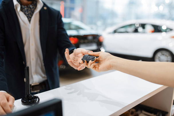 Man takes the key from new auto in car dealership. Customer and saleswoman in vehicle showroom, male person buying transport, automobile dealer business - Foto, immagini