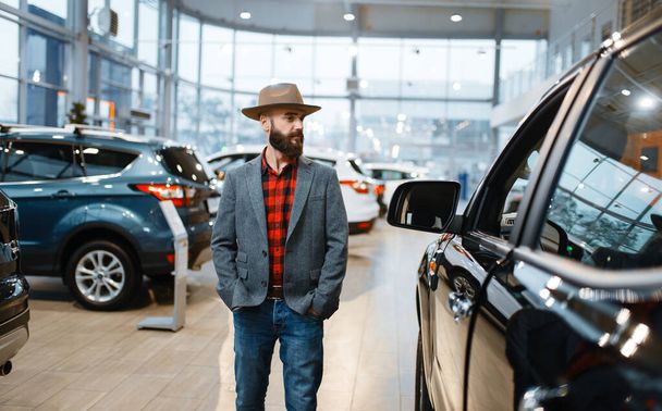Man in a cowboy hat choosing new automobile in car dealership. Customer in vehicle showroom, male person buying transport, auto dealer business - Foto, Imagen