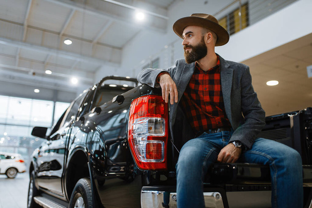 Muž v klobouku pózuje v novém pickupu v prodejně aut. Zákazník ve vozidle showroom, muž osoba kupující dopravu, auto dealer obchod - Fotografie, Obrázek