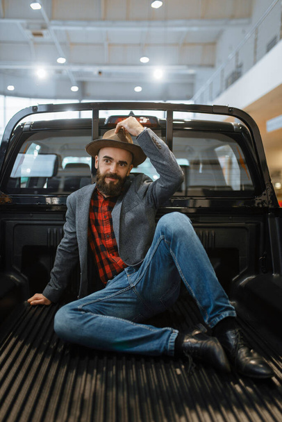 Homem sorridente posa na parte de trás do caminhão novo pick-up na concessionária de carros. Cliente no showroom do veículo, pessoa masculina que compra o transporte, negócio do negociante do automóvel
 - Foto, Imagem