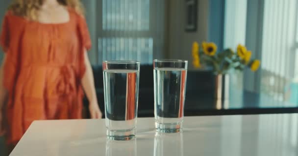 A young woman wearing a red dress is approaching a kitchen counter and picks up two glasses of water. Panning shot in slow motion. - Кадры, видео