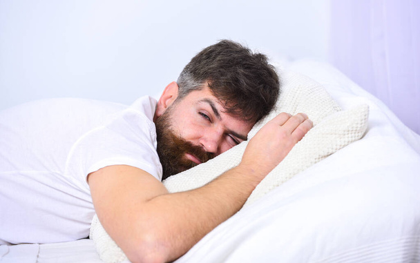 Man in shirt laying on bed, white wall on background. Macho with beard and mustache sleeping, relaxing, having nap, rest. Guy on tired face sleeping on white sheets and pillow. Nap and siesta concept. - Foto, Bild
