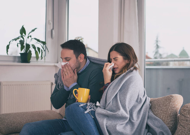 Young couple sitting on sofa covered with blanket, sneezing and blowing nose. Cold and flu season  - Foto, Imagem