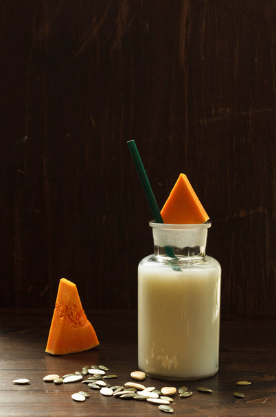 Portion pflanzliche Milch ohne tierische Fette aus Kürbiskernen in einer Glasflasche und Kürbisscheiben auf dunklem Hintergrund mit Platz - Foto, Bild