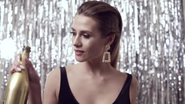 Portret van prachtige elegante sensuele blonde vrouw in de mode zwarte jurk met een fles champagne op tinsel achtergrond in studio - Video