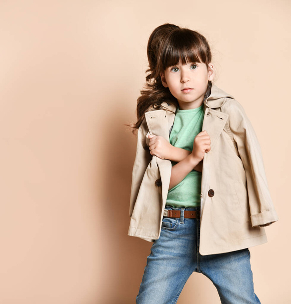 a little brunette girl in a beige raincoat, with her arms crossed over her chest - Foto, afbeelding
