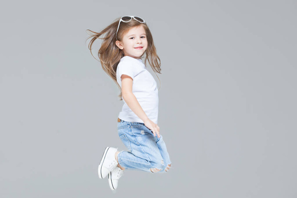 Kid girl preschooler in blue jeans, white t-shirt and sunglasses is jumping isolated on grey background - Photo, Image
