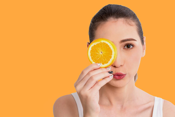 Portret pięknej, radosnej młodej, uroczej, szczęśliwej Azjatki trzymającej soczysty plasterek pomarańczowego owocu ukrywający oko za i patrzący na odizolowaną kamerę na pomarańczowym tle.Zdrowe jedzenie koncept.Diet - Zdjęcie, obraz
