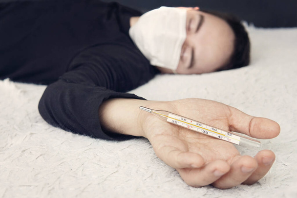 ziek persoon in het dragen van een beschermende medische masker in bed ligt op het bed en houdt een thermometer met een hoge temperatuur in zijn hand. Thermometer close-up. Man met acute mannelijke verkoudheid meet koorts - Foto, afbeelding
