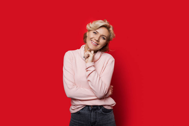Charming caucasian lady with blonde hair is looking and smiling at camera while posing on a red wall - Photo, Image