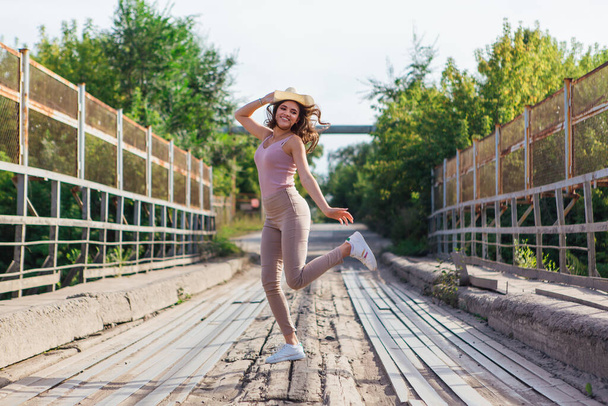 Warm summer portrait of a young beautiful woman wearing cowboy hat jumping on the old rusty bridge during sunset. - Valokuva, kuva