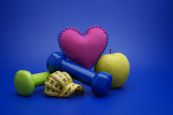 Still life with small dumbbells, measuring tape and green apple, as a concept of healthy nutrition and sports diet in close-up on blue background - Photo, Image