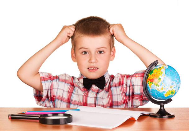 Befuddled boy sitting at a table on a notebook, pencils, magnifying glass and globe isolated on white background. - Фото, изображение