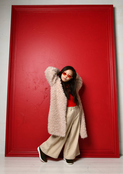 Cool asian kid girl in furcoat and wide pants is posing with her hands up and behind her head - Foto, Imagem