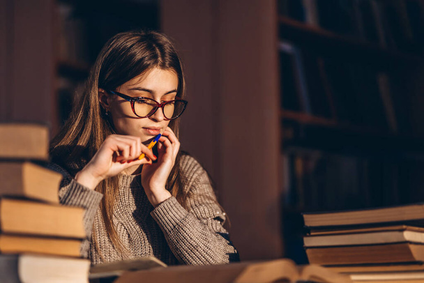 Young student in glasses preparing for the exam. Girl in the evening sits at a table in the library with a pile of books. - Foto, Imagen