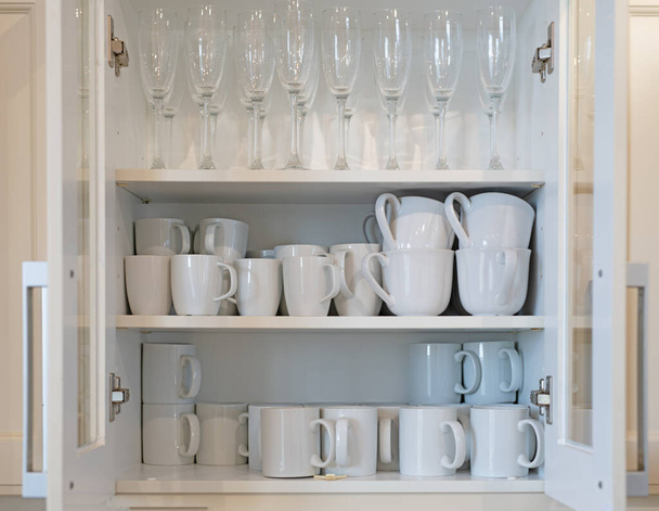 Kitchen cupboard with the door open and full of white mugs and champagne glasses - Fotó, kép