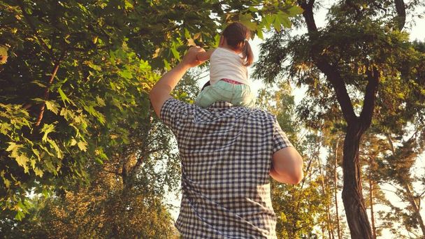 Papa trägt sein geliebtes Kind auf den Schultern im Park. Der Vater geht mit seiner Tochter auf den Schultern unter Bäumen. Kind mit Eltern geht am freien Tag spazieren. Glückliche Familie entspannt im Park. - Foto, Bild