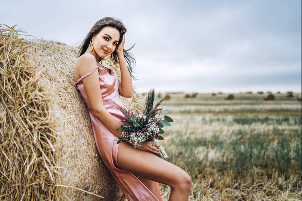 Young brunette in a pink satin dress, without linen standing near hay bales in windy weather. A woman holds a bouquet of wildflowers. - Fotografie, Obrázek