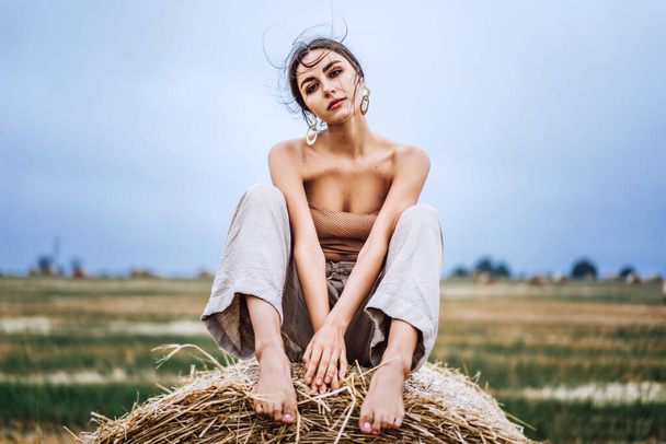 Brunette in linen pants and bare shoulders sitting on a hay bales in warm autumn day. Woman looking at camera. Behind her is a wheat field. - Fotó, kép
