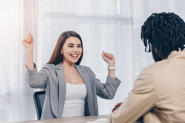 Selective focus of happy and excited employee with african american recruiter at job interview in office - Фото, изображение
