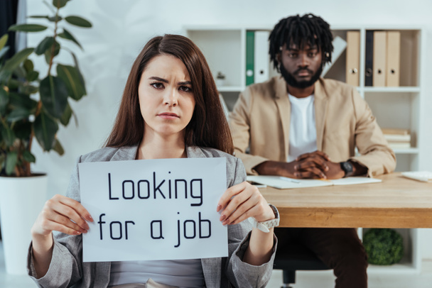 Worried employee showing placard with looking for a job lettering and african american recruiter in office - Photo, Image