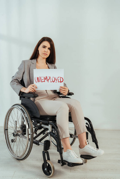 Disabled woman looking at camera and holding placard with unemployed lettering on wheelchair on white  - Foto, afbeelding